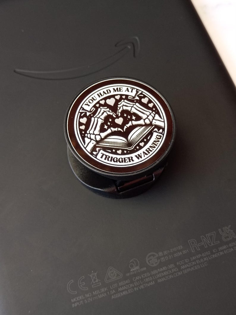 May include: A black and brown phone grip with the text "YOU HAD ME AT TRIGGER WARNING" in a circular design. The design includes skeleton hands, hearts, and an open book. The grip is attached to a black electronic device.