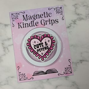 May include: A pink and white magnetic Kindle grip with the words "Cute & Psycho" on a heart-shaped design. The grip is on a pink and white card with the words "Magnetic Kindle Grips".