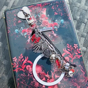 May include: A decorative keychain with multiple charms, including a red bow, skeleton hand, black rose, skull, and an owl. The keychain is attached to a book cover with a floral design and the text "I LOVE BOOKS".