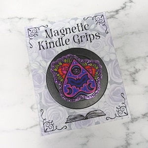 May include: A black magnetic Kindle grip featuring a purple glitter planchette topper with a bat silhouette, crescent moons, and red floral accents. The product is mounted on a display card with the text Magnetic Kindle Grips and a book icon.