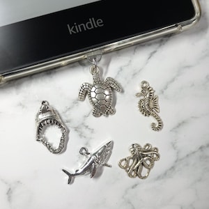 May include: A selection of silver-coloured ocean-themed charms, including a turtle, seahorse, shark, shark jaw, and octopus. The charms are displayed near a Kindle e-reader. The background is a white and grey marble pattern.