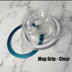 May include: Clear plastic Mag Grip with a white ring and a teal accent. The text "Mag Grip - Clear" is printed in black. The object is on a white and gray marble surface.