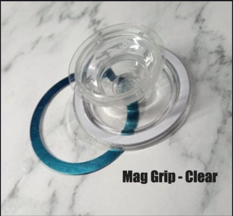 May include: Clear plastic Mag Grip with a white ring and a teal accent. The text "Mag Grip - Clear" is printed in black. The object is on a white and gray marble surface.