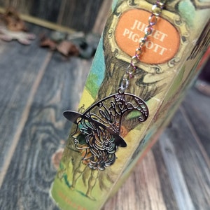 May include: A silver bookmark featuring a detailed cutout of a witch wearing a hat with the word "Witch" inscribed on it. The bookmark is attached to a book with the title "Juliet Pigott" on the cover.