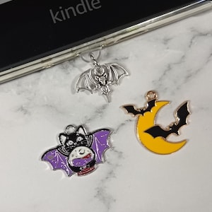 May include: Three Halloween charms. One is a silver bat with outstretched wings. Another is a black and purple bat with a crystal ball. The third is a yellow crescent moon with a black bat silhouette. The word "kindle" is visible.