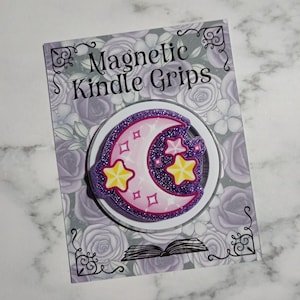 May include: A magnetic Kindle grip with a purple glitter crescent moon and yellow stars design. The grip is on a card with the text "Magnetic Kindle Grips" and a floral background.