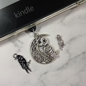 May include: A silver owl pendant on a black cord, with a crescent moon design and a smaller owl charm. A black raven charm is also present. The word "kindle" is visible on a black electronic device.