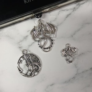 May include: Three silver dragon pendants of different sizes. The largest dragon has outstretched wings and a curled tail. The other two pendants feature detailed dragon designs. The word "kindle" is visible in the background.