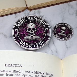 May include: Two round stickers with a dark romance book club theme. The larger sticker features a skull with roses and a book, while the smaller sticker features a similar design with a smaller skull and book. Both stickers have a black background with white text and a purple design.