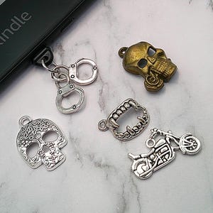 May include: A collection of metal charms, including a skull, handcuffs, motorcycle, and vampire fangs. The skull charms are in silver and gold tones. The word "kindle" is visible on a black electronic device.