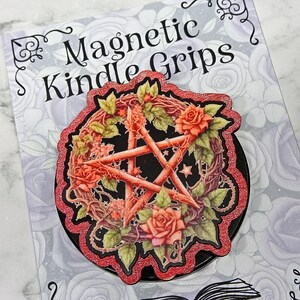 May include: A black magnetic Kindle grip featuring a red pentagram design with roses and leaves. The grip has a glittery red border and is set against a floral background. The text "Magnetic Kindle Grips" is visible.