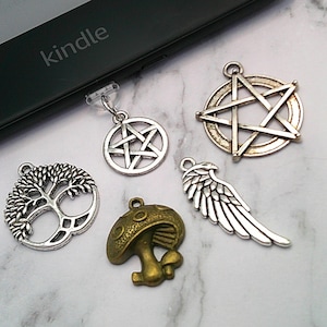 May include: A selection of silver and bronze charms, featuring a pentagram, a tree of life, a mushroom, and a wing. The word "kindle" is visible on a black electronic device.