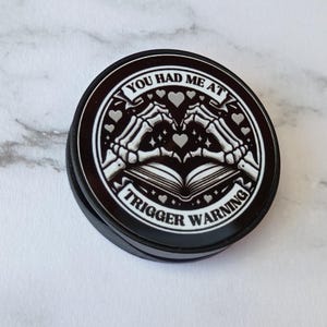 May include: Black round pop-up phone grip with a white graphic. The graphic features skeleton hands forming a heart over an open book, with the text "YOU HAD ME AT TRIGGER WARNING" in a circular design.