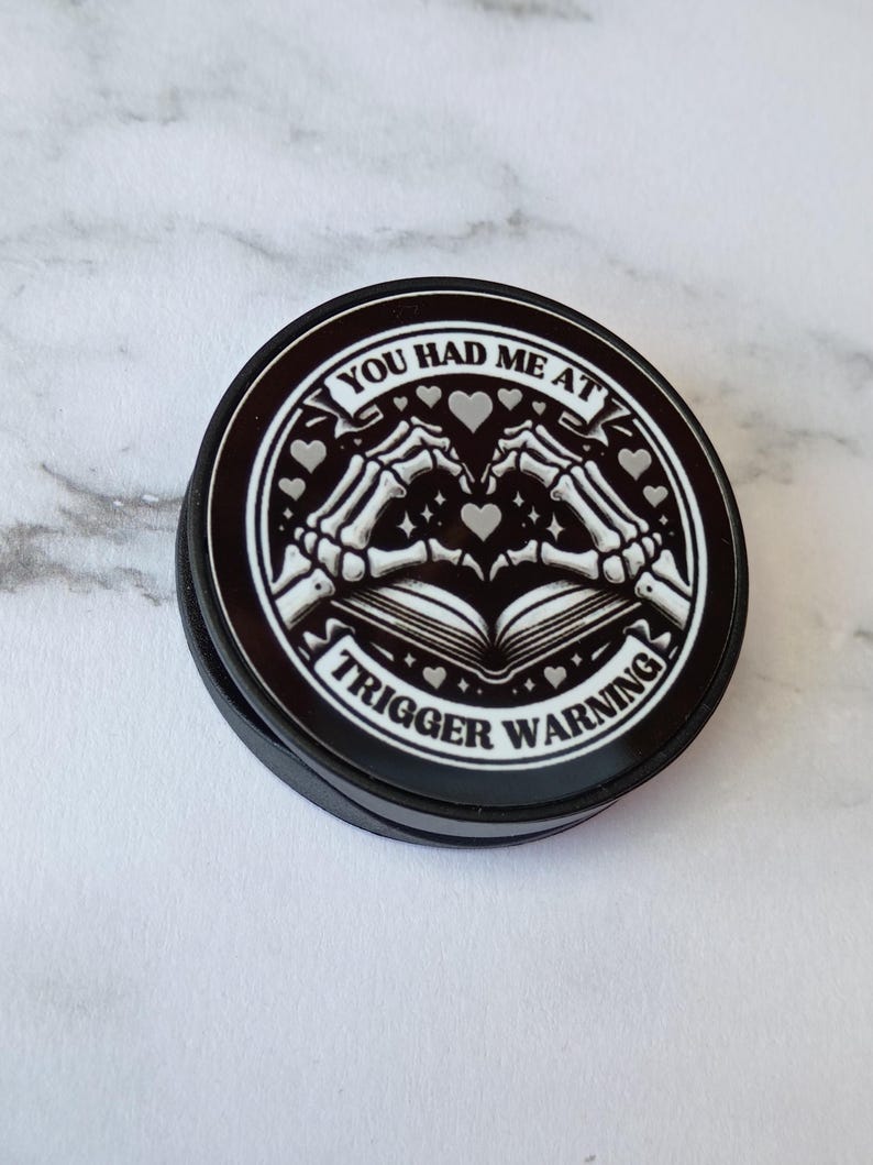 May include: Black round pop-up phone grip with a white graphic. The graphic features skeleton hands forming a heart over an open book, with the text "YOU HAD ME AT TRIGGER WARNING" in a circular design.