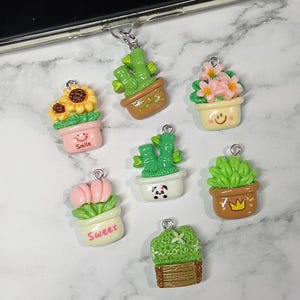 May include: A selection of miniature plant charms, each in a small pot. The charms feature sunflowers, cacti, and flowering plants in various colours. Some charms have the words "Smile" and "Sweet" on them.
