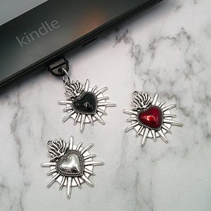 May include: Three heart-shaped pendants with a sunburst design. One pendant has a black heart, another a red heart, and the third a silver heart. The word "kindle" is visible on a black electronic device in the background.