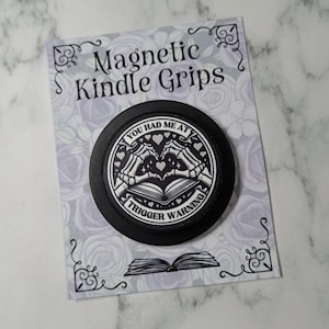 May include: A black magnetic Kindle grip featuring the text "YOU HAD ME AT TRIGGER WARNING" and a graphic of skeleton hands forming a heart over an open book. The grip is on a decorative card with the words "Magnetic Kindle Grips".