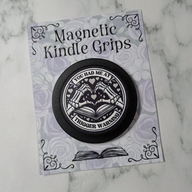 May include: A black magnetic Kindle grip with the text "YOU HAD ME AT TRIGGER WARNING" and a graphic of skeleton hands forming a heart over an open book. The grip is on a decorative card with the words "Magnetic Kindle Grips".