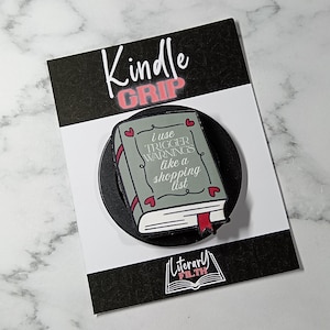 May include: A circular black phone grip featuring a rectangular book-shaped topper. The book is sage green with red hearts and the text: I USE TRIGGER WARNINGS like a shopping list. The backing card displays the text Kindle GRIP and Literary FILTH.