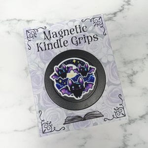 May include: A black magnetic Kindle grip featuring a bat design. The design shows three bats with purple wings, set against a starry night sky with a crescent moon. The packaging reads "Magnetic Kindle Grips".
