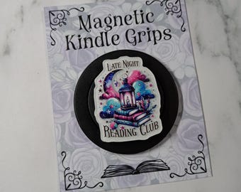 May include: A black circular magnetic Kindle grip featuring a sticker with a lantern, stacked books, and clouds in shades of pink, blue, and purple. The text reads Late Night Reading Club. It is mounted on a card labeled Magnetic Kindle Grips.