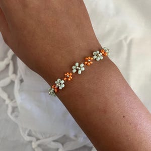 Pode incluir: Uma pulseira de contas com um design floral. A pulseira tem pequenas contas em forma de flor, verde claro e laranja, enfiadas. A pulseira &eacute; usada em um pulso contra um fundo branco.