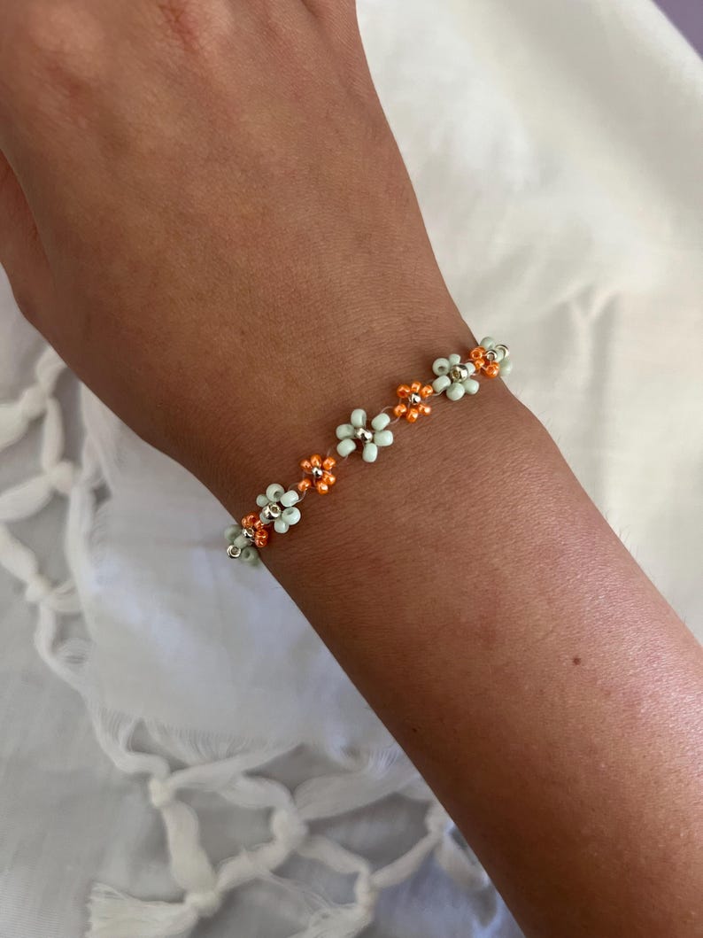 Pode incluir: Uma pulseira de contas com um design floral. A pulseira tem pequenas contas em forma de flor, verde claro e laranja, enfiadas. A pulseira &eacute; usada em um pulso contra um fundo branco.