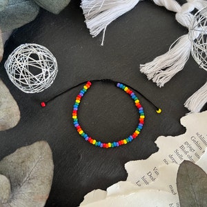 Könnte beinhalten: Ein Regenbogen-Perlenarmband mit schwarzem Band. Das Armband besteht aus kleinen, bunten Perlen und ist mit einem Knoten gebunden.