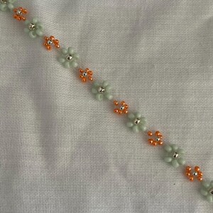 Pode incluir: Uma pulseira de contas com um design floral. A pulseira tem contas de flores verde claro com centros prateados e contas de flores laranja. &Eacute; exibida em um fundo de tecido branco.