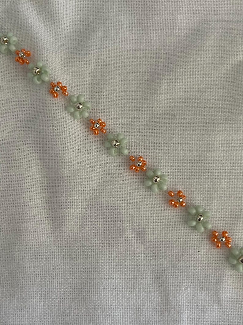 Pode incluir: Uma pulseira de contas com um design floral. A pulseira tem contas de flores verde claro com centros prateados e contas de flores laranja. &Eacute; exibida em um fundo de tecido branco.