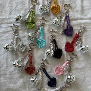 Puede incluir: Una colección de llaveros con dijes de bolas de discoteca, corazones de ganchillo y detalles en forma de estrella. Cada llavero tiene un corazón de diferente color y una palabra con cuentas, como "FEARLESS" o "EVERMORE". Los llaveros están sobre una superficie blanca.