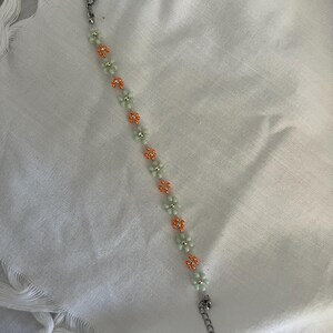 Pode incluir: Uma pulseira de contas com um design floral. A pulseira alterna contas em forma de flor verde claro e laranja. Possui um fecho e uma corrente prateados.