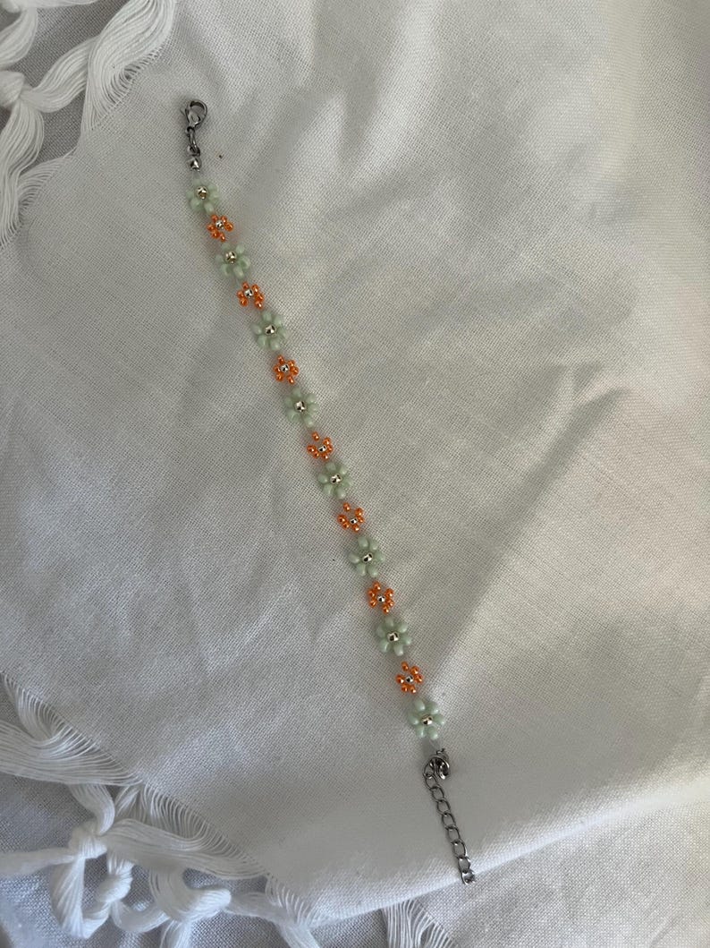 Pode incluir: Uma pulseira de contas com um design floral. A pulseira alterna contas em forma de flor verde claro e laranja. Possui um fecho e uma corrente prateados.