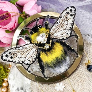 May include: A gold-framed, circular ornament featuring a detailed embroidered bumblebee with black and yellow stripes, white fur, and clear crystal accents. The ornament hangs from three gold chains with black beads.