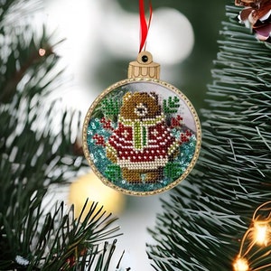 May include: A round Christmas ornament with a beaded bear design. The bear wears a red sweater with white accents and a green scarf. The ornament has a gold-colored frame and a red ribbon for hanging.