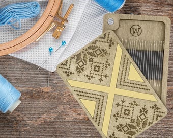 May include: A wooden embroidery hoop, blue thread spools, and a needle case with needles are arranged on a wooden surface. The needle case has a geometric design. Embroidery pins and a piece of white fabric are also present.