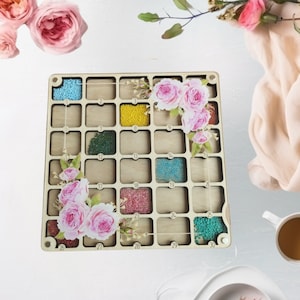 Könnte beinhalten: Ein Holzperlen-Organizer mit 30 nummerierten Fächern, gefüllt mit bunten Perlen. Der Organizer ist mit rosa Rosenblüten verziert. Eine Tasse Kaffee und Blumen befinden sich im Hintergrund.