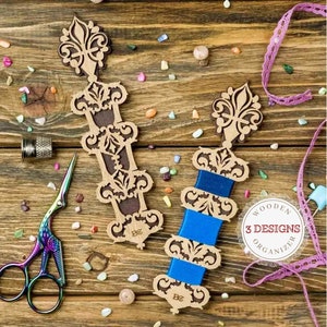Pode incluir: Dois organizadores de fita de madeira com designs intrincados e bobinas de fita azul. Os organizadores estão sobre uma superfície de madeira com contas coloridas e renda. O texto "WOODEN 3 DESIGNS ORGANIZER" é visível na imagem.