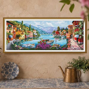 Könnte beinhalten: Ein rechteckiger gerahmter Wandkunstdruck, der eine mediterrane Küstendorf-Szene mit farbenfrohen Gebäuden, kleinen Booten auf blauem Wasser, leuchtend violetten und rosa Blumen im Vordergrund und entfernten Bergen unter einem hellen Himmel zeigt.