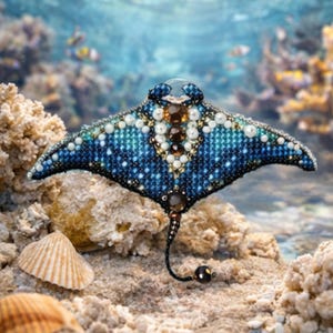 May include: A handcrafted, beaded stingray brooch. The brooch features a blue and white beaded design with brown and clear accents. The stingray has a long, black tail with a dark bead at the end. The background shows coral and seashells.