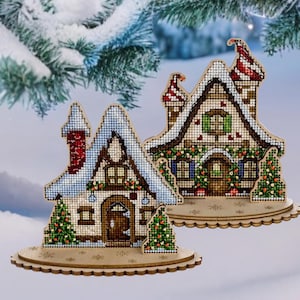 以下が含まれることがあります： 刺繍の細かいディテールが施された木製のクリスマスハウス2つ。雪、緑、赤いアクセントで飾られています。家は木製の台座の上に置かれています。