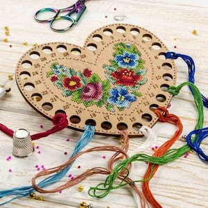 May include: A heart-shaped wooden thread organiser with 29 numbered holes for storing embroidery floss. The organiser is decorated with a colourful floral embroidery design. Several strands of floss are threaded through the holes.