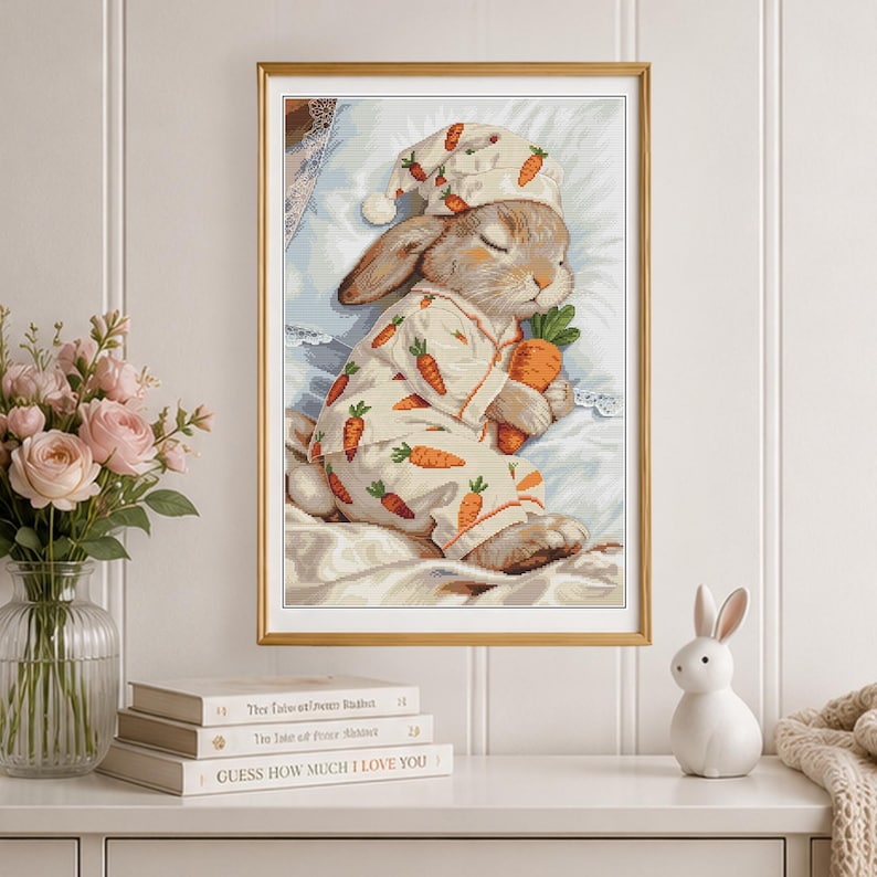 Peut inclure: Une impression d'art encadr&eacute;e repr&eacute;sentant un lapin endormi portant un pyjama et un bonnet de nuit d&eacute;cor&eacute;s d'un motif de carottes. Le lapin repose sur un oreiller blanc et tient une grosse carotte orange. L'&oelig;uvre est r&eacute;alis&eacute;e dans des tons pastel.