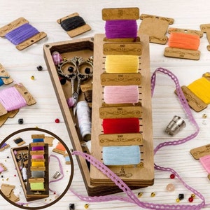 May include: A wooden box with a variety of colorful threads and a small sewing kit. The box has a hinged lid and is decorated with a floral design. The threads are arranged in a vertical row and are labeled with numbers. The sewing kit includes a pair of scissors, a needle threader, and a thimble.