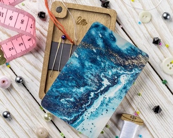 May include: A wooden box with a blue and gold marbled design on the lid. The box contains sewing needles and a small piece of fabric. The box is surrounded by beads and other sewing supplies.