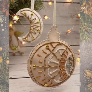 Könnte beinhalten: Zwei handgefertigte Weihnachtsschmuckstücke. Eines hat die Form eines Halbmonds, das andere ist kugelförmig. Beide sind cremefarben mit goldenen und weißen Perlenverzierungen und dekorativen Nähten. Sie hängen an weißen Bändern.