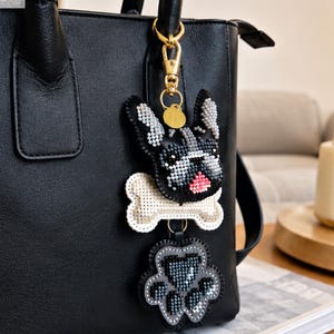 May include: A black leather handbag featuring a beaded keychain charm. The charm consists of a black and white French bulldog head, a white bone, and a gray paw print, all connected by a gold-toned metal lobster clasp and a small circular tag.