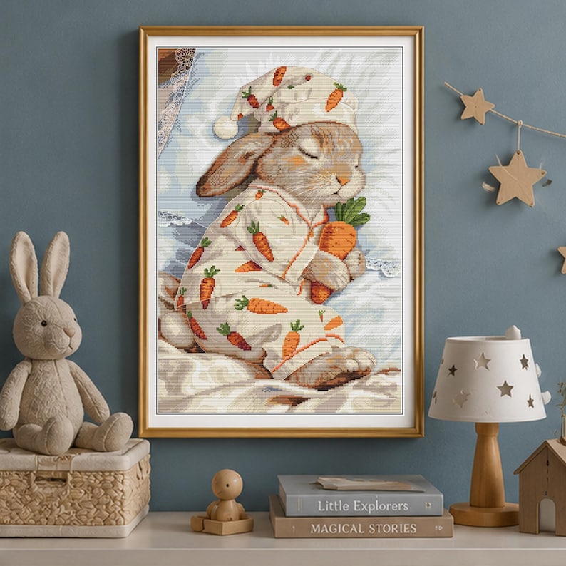 Peut inclure: Une &oelig;uvre au point de croix encadr&eacute;e repr&eacute;sentant un lapin endormi portant un pyjama et un bonnet de nuit &agrave; motifs de carottes orange. Le lapin tient une grosse carotte orange tout en se reposant sur un oreiller blanc dans un cadre dor&eacute;.
