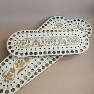 Könnte beinhalten: Mehrere unfertige ovale Taschenböden aus Holz mit floralen Mustern und nummerierten Löchern. Die Böden sind hell und haben eingravierte Blumenmuster und gleichmäßig verteilte Löcher.