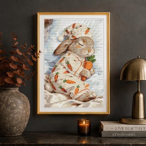 Peut inclure: Une impression d'art encadr&eacute;e repr&eacute;sentant un lapin endormi portant un bonnet de nuit et un pyjama cr&egrave;me d&eacute;cor&eacute;s d'un motif de carottes orange. Le lapin tient une grosse carotte orange tout en se reposant sur un oreiller et une couverture blancs.